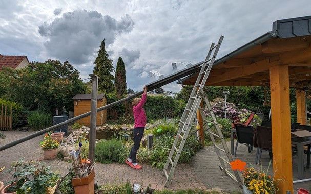 Auf einer langen Leiter, die sehr schräg vom Erdboden bis auf das Dach vom Gartenpavillon liegt, liegt eine 250W-Solarzelle mit angebauter Aufständerung. Die Solarzelle wird auf der Leiter von unten aufs Dach geschoben.