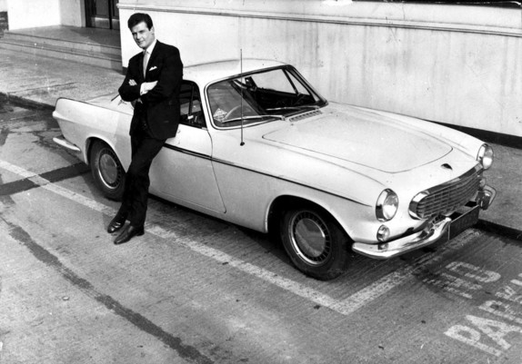 Roger Moore leans against a white or cream Volvo P1800 coupe, possibly a 1962, in a publicity photo for his TV show “The Saint”