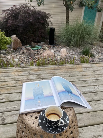 A hardcover graphic novel and a now-empty coffee cup and saucer rest precariously on a wicker side table which itself is on a wooden deck facing a stone garden featuring Japanese Maples, a basalt column water fountain, a bird bath, and grasses. Behind the garden is a wall covered in siding with a teal door facing a path.