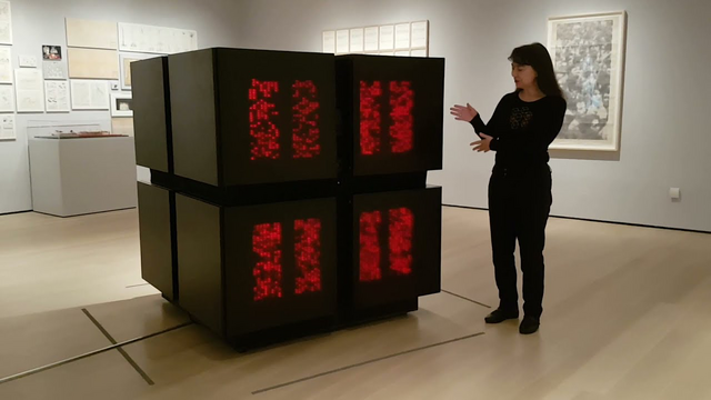 Industrial Designer Tamiko Thiel gestures towards a Connection Machine. It appears to be a human-scale collection of four black cubes assembled into a larger cube. On the front of each cube facing us are two arrays of red lights that individually turn on and off rapidly when the system is in use.